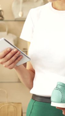 Parlak beyaz tişörtlü ve son moda yeşil pantolonlu genç bir kadın akıllı telefonuyla yakın plan fotoğraf çekerken büyüleyici bir turkuaz ayakkabıya hayran kalıyor. Zarif aksesuarlarla çevrili, modern alışverişin heyecanını yayıyor.