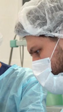 Yüksek bahisli bir ameliyathanede steril mavi bir önlük ve maske giymiş odaklanmış bir erkek cerrah ameliyatın karmaşıklığını ustalıkla incelerken özenli bayan asistanı titizlikle dolgu yapıyor. Bu sürükleyici sahne,