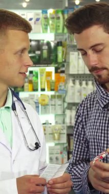 Kendini işine adamış beyaz önlüklü bir doktor elinde bir şişe hap tutan meraklı bir erkek hastaya ilaç talimatlarını özenle açıklar. Renkli sağlık ürünleriyle dolu enerjik eczane raflarına karşı kurulmuş bu ilgi çekici sahne...