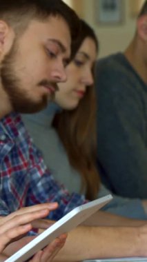 Kendinden emin beyaz bir kadın tabletiyle meşgul oluyor, esmer saçı kameraya doğru dönerken yüzünü çerçeveliyor. Arka planda, odaklanmış sınıf arkadaşları, sakallı bir genç adam, özenle not alır, dinamik ve canlı bir şekilde