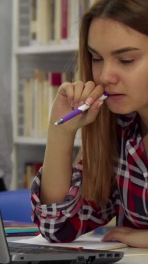 Kararlı bir beyaz kız öğrenci, canlı bir kütüphanede çalışırken dizüstü bilgisayarına dalmış kalemini ısırıyor. Yüksek kitap raflarıyla çevrelenmiş olan kareli gömleği, onu yansıtan sakin atmosfere biraz renk katıyor.