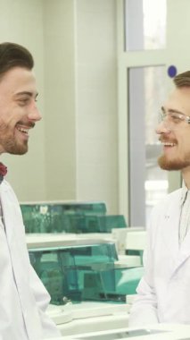Parlak, güneşli bir laboratuvarda, iki genç erkek asistan neşeli bir sohbete girişiyor, yüzleri neşe ve arkadaşlıkla parlıyor. El değmemiş laboratuvar önlükleri ve koruyucu gözlük giyip takım çalışmasının enerjisini temsil ediyorlar.