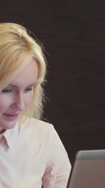 Kararlı sarışın bir kadın parlak, ahşap bir masada oturuyor, laptopuyla derinden meşgul, sıcaklık ve pozitiflik saçıyor. Zengin ahşap tonlar ve yumuşak ışıklandırma ile vurgulanmış rahat ofis atmosferi, onun profesyonel ruhunu tamamlıyor.