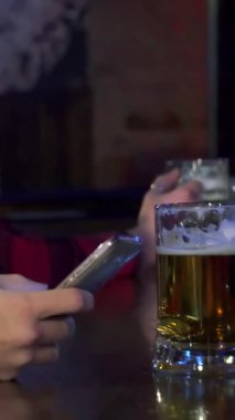 Şık sakallı çekici bir genç adam akıllı telefonuyla derinden meşgul. Etrafı hareketli bir barın enerjisiyle çevrili. Sıcak ortam aydınlatması, rahatlatıcı birasını yudumlarken rahat tavırlarının altını çizerek hoş bir parıltı yayıyor.