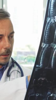 El değmemiş beyaz önlüklü genç bir erkek doktor, modern tıbbi ekipmanlarla dolu güneşli ofisinde röntgenini incelerken pozitiflik yayıyor. Geniş pencerelerden süzülen doğal ışıkla güçlendirilmiş davetkar atmosfer,
