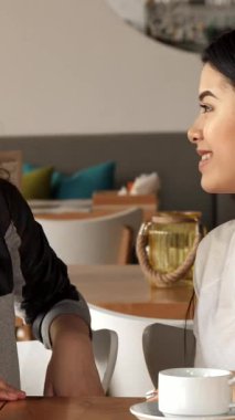 Canlı bir kafe ortamında, iki neşeli kız, bir Asyalı ve bir Kafkasyalı, elleriyle kalp şekilleri yaratıyorlar, dostluk ve neşeyi somutlaştırıyorlar. Süslü dekorlar ve beyaz kahve fincanlarıyla çevrelenmiş kahkahaları havayı dolduruyor, enfes bir görüntü yakalıyor.