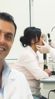 Sıcak kalpli bir doktor, ön planda hoş bir gülümsemeyle kendine güven ve yakınlık yayıyor. Arka planda, kendini işine adamış bir göz doktoru titizlikle bir hastanın gözlerini inceleyerek, bir bakıcı ve profesyonel atmosfer yaratıyor.