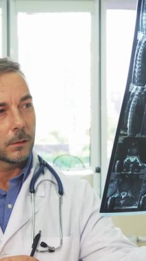 Gıcır gıcır beyaz laboratuvar önlüğü giymiş odaklanmış genç bir adam X-Ray görüntüsünü yüksek tutuyor ve parlak, modern ofisinde ciddi bir ifadeyle inceliyor. Boynundaki steteskop ve pencerelerden süzülen yumuşak doğal ışık...