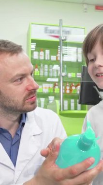 Kendini işine adamış beyaz önlüklü bir erkek eczacı heyecanlı küçük bir çocukla sıcak etkileşime girer. O da hevesle parlak bir turkuaz şampuan şişesini inceliyor. Bu hoş sahne sıcaklığı ve ilgiyi yansıtıyor. Renkli bir eczaneyi sergiliyor.