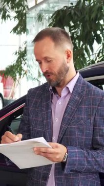 Gösterişli, modern bir araba galerisinde, verimli yeşilliklerle süslenmiş, keyifli bir alıcı ve cilalı bir satıcı, araba alımlarını neşeyle sonuçlandırmak için kontratı imzalarken ışıl ışıl gülümserler. Şık mavi ekose ceketli adam coşkulu görünüyor.