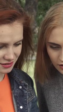 Yeşilliklerle dolu yemyeşil bir parkta, biri parlak kızıl saçlı, diğeri sarı bukleli iki genç kadın, bir akıllı telefonu neşeyle paylaşıyor. Onların bulaşıcı kahkahaları ve parlak gülümsemeleri, canlı bir dostluk ortamı yaratıyor.