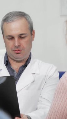 Şefkatli bir erkek doktor, yaşlı bir kadın hastayla düşünceli bir konuşma yaparken röntgen taramalarını inceliyor ve sıcak ve destekleyici bir sağlık ortamı yaratıyor. Bu sahne, tıbbi araştırmalar arasındaki hayati bağlantıyı vurguluyor.