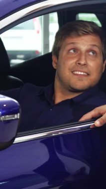 Kısa saçlı neşeli bir adam şık mavi arabasında oturur ve onun etkileyici özellikleri hakkındaki görüşlerini hevesle paylaşan satıcıyla ilgilenir. Canlı sohbetleri heyecan saçıyor, dinamik bir atmosfer yaratıyor.