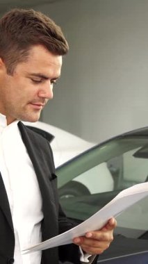 Düzgün kesilmiş sakallı neşeli bir adam yeni arabasının anahtarlarını gururla sergilerken şık, modern araçlarla çevrili şık bir galeride mutluluk saçıyor. Sivri siyah takım elbise ve beyaz gömlek giymişti. Kendine güveni tamdı.