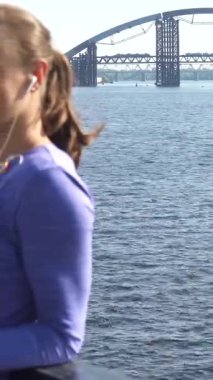 Büyüleyici bir sahnede, uzun, siyah saçlı, at kuyruklu genç bir kadın ışıldayan nehrin kenarındaki korkulukta dinleniyor. Canlı bir menekşe bluz giyer, kulaklıklarıyla dikkatle dinler, sükuneti somutlaştırır ve odaklanır.