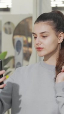 Neşeli genç bir kadın şık alışveriş merkezinin ortasında elinde şık alışveriş torbalarıyla akıllı telefonuyla neşeyle selfie çekiyor. Bu canlı sahne mutluluk saçıyor ve moda kıyafetlerini sergileyerek perakende terapinin heyecanını simgeliyor.