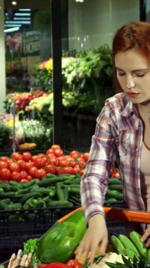 Canlı bir süpermarkette alışveriş arabasında renkli bir dizi taze meyve ve sebze organize ederken canlı kızıl saçlı neşeli bir beyaz kadın. Olgun domateslerden, salatalıklardan ve yemyeşil çiçeklerden oluşan bir kaleydoskopla çevrili.