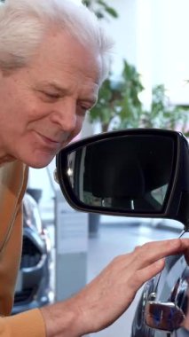 Gümüş saçlı seçkin bir kıdemli adam modern bir bayilikte çarpıcı bir arabanın parlak kanadını yakından incelemek için öne eğiliyor. Düşünceli ifadesi ve nazik dokunuşu takdire şayan, parlak, davetkar bir şekilde araçların zarafetini vurguluyor