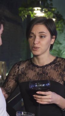 Siyah dantelli elbisesi olan çarpıcı genç bir kadın, her ikisi de yemyeşil süslerle bezenmiş şık bir barda neşe saçan yakışıklı erkek arkadaşıyla hoş bir konuşma yapıyor. Yumuşak ışıklandırma ve zarif dekorla vurgulanan samimi atmosfer,