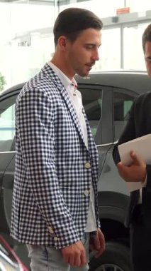 Şık bir araba galerisinde, ciddi bir satıcı sözleşmeyi sunarken, şık ve kareli ceket giymiş bir alıcı ayrıntıları yakından inceler. Odaklanmış bir tartışmaya girerken atmosfer heyecan vericidir.