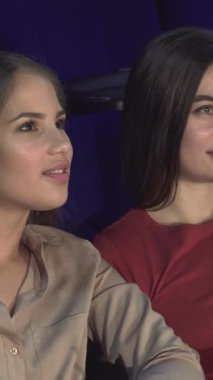 İki genç kadın, biri yumuşak bej bir bluz ve diğeri canlı kırmızı bir bluz içinde, samimi bir sinemada yan yana oturuyorlar, yüzleri heyecanlı bir fısıltıyı paylaşırken parlıyor. Bu büyüleyici an, kahkaha ve neşeyle dolu, güzel bir şekilde