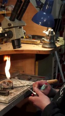 Yetenekli genç beyaz bir kuyumcu, bir gaz lambasını ustalıkla tutuşturuyor. Güç topuzuna ince ayar çekerken alevler yanıp sönüyor. Bu dinamik sahne mücevher yapımının özünü yakalar, bir dizi alet ve malzemeyle çevrili,