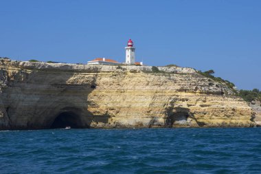 A view of Alfanzina Lighthouse above the rugged limestone cliffs and grottos along the Algarve coast in Portugal. clipart