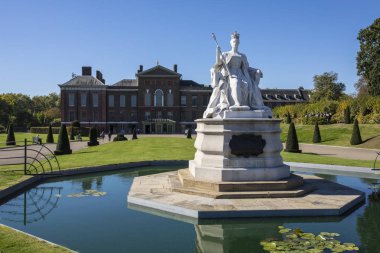 Londra, İngiltere - 27th Eylül 2018: Kensington Gardens Londra'da Kraliçe Victoria taç giyme elbisesi 1837 yılında tasvir eden bir heykel. Kensington Sarayı arka planda olduğunu.