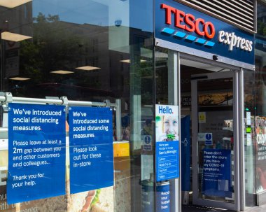 Londra, İngiltere - 24 Haziran 2020: Londra 'da bir Tesco Express süpermarketinin vitrinindeki işaretler, müşterilere COVID-19' un yayılmasını önlemek için sosyal mesafeleme tedbirleri uyguladıklarını bildiriyor.