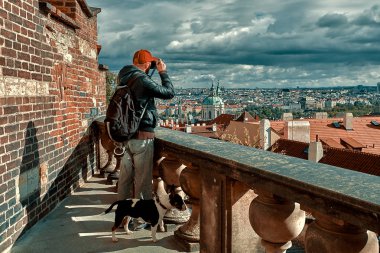 Young man with a backpack and a puppy dog is taking aerial view picture photo of the Old Town Prague, Czech Republic. Touristic travel photo conception clipart