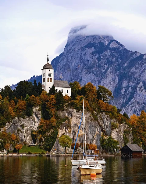 Schöner malerischer Sonnenuntergang über den österreichischen Alpen. Boote, Yachten im Sonnenlicht vor der Kirche auf dem Felsen mit Wolken über dem Traunstein am Alpensee bei Hallstatt salzkammergut Österreich Stockbild