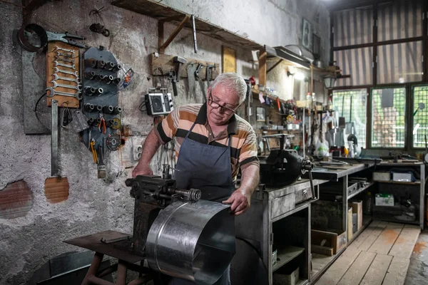 An older man working with sheet metal in his workshop. A worker works carefully with sheet metal