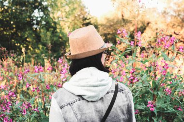 Hipster bir kız arkası seyirciye dönük olarak yeşillik ve kır çiçeklerinin arka planında duruyor. Gençlik tarzı. Genç bir kadın orkideye benzeyen pembe, fuşya renkli çiçeklere bakar. Yaz akşamı, gün batımında, ormanın kenarındaki tarlada yürü..