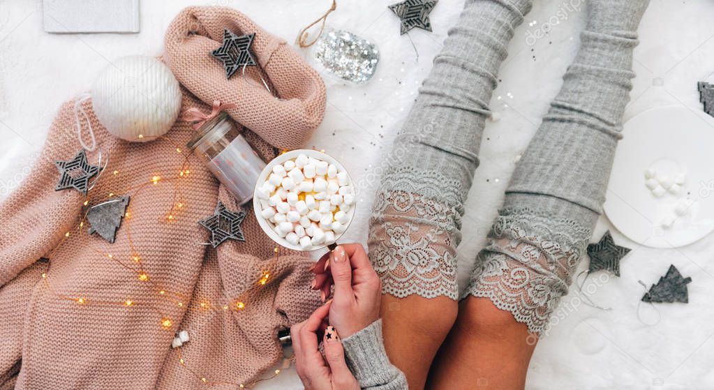 Lifestyle composition. Cozy winter,, warm woollen socks. Woman on white blanket with marshmallow cup and christmas decorations. Holiday relax.  