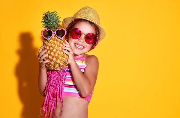 滑稽愉快的女孩在泳装 粉红色的眼镜和帽子与菠萝是笑在颜色黄色背景 — 图库照片
