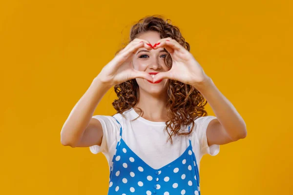 Beautiful funny girl holds hands in shape of a heart on yellow b — Stock Photo, Image