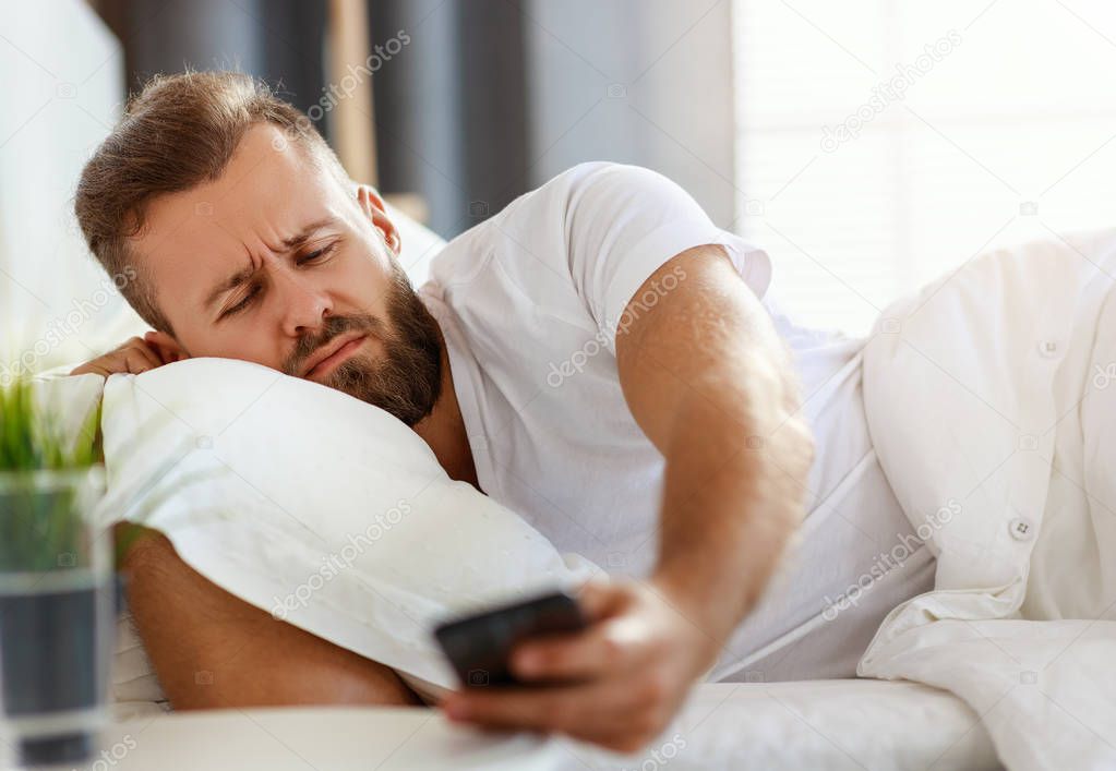 young bearded man wakes up in   morning in his bed with phone al