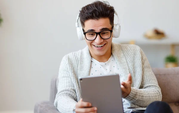 在舒适的客厅里 坐在沙发上 戴着耳机和眼镜的积极的年轻男性在平板电脑上通过视频链接与朋友聊天 — 图库照片