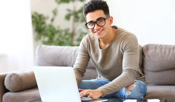 身穿休闲装 戴着眼镜的积极向上的年轻人一边坐在沙发上 一边微笑着拍照 一边浏览笔记本电脑上的数据 并在网上做远程项目 — 图库照片