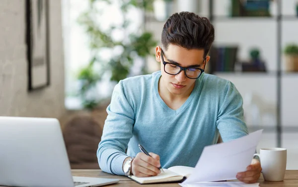 Ethnic young man  reading document and writing in planner while sitting at table and working on remote project at hom