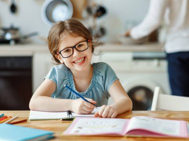 Gözlüklü, çalışkan bir ilkokul öğrencisi kameraya bakıp gülümserken evde uzaktan çalışmalar sırasında, tanınmayan bir anneyle angarya bir evde yemek hazırlarken ve fotoğraf makinesi kullanırken.
