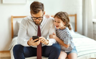 Serious frowned busy man in formal wear and eyeglasses reading business news on smartphone while cheerful little daughter sitting nearby and asking for attention during morning in bedroom at hom clipart