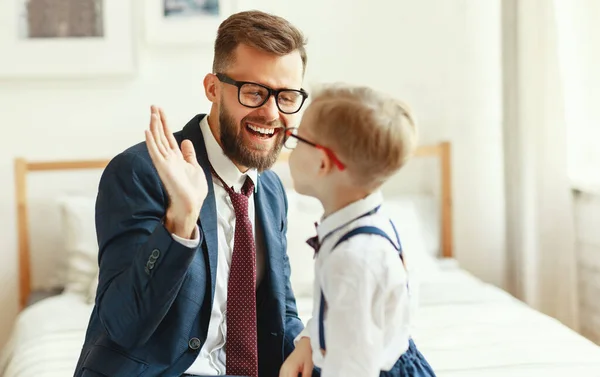 学校での成功を祝いながら ベッドの上に座って 小さな息子に5を与える正式な衣装や眼鏡で陽気な若いひげを生やしたビジネスマン — ストック写真