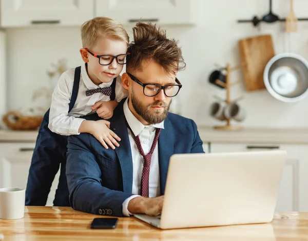 奇妙な小さな息子はノートパソコンの画面を見ている間忙しいお父さんで正式な衣装でしくじった髪で家でリモートビジネスプロジェクトに取り組んで — ストック写真