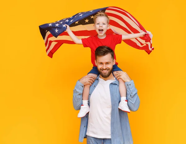 黄色の背もたれに対する独立記念日を祝いながら 肩にアメリカの旗で興奮した男の子を運ぶ幸せな髭の男 — ストック写真