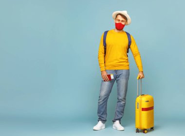 Koruyucu maskeli adam bavulun yanında duruyor ve salgın sırasında Blue Backdro 'ya karşı tatile çıkarken kameraya bakıyor.