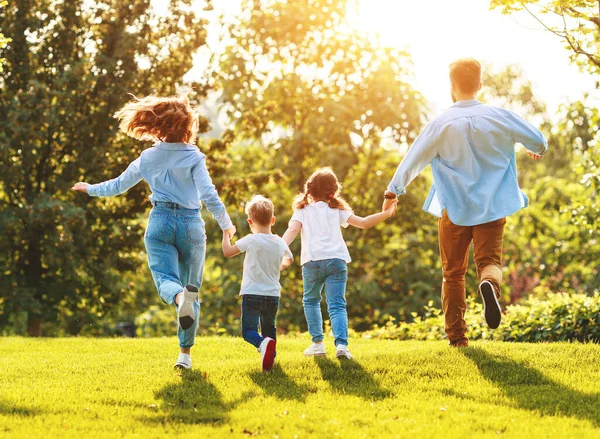 夏の緑の芝生で一緒に時間を楽しみながら手をつないで走るカジュアルな服を着た子供たちを持つ認識できない幸せな両親Da — ストック写真