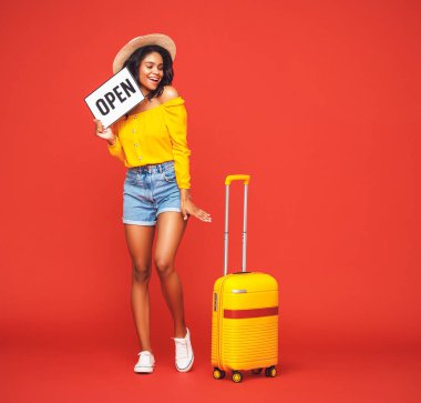 concept of travel. happy excited young black   optimistic ethnic    female tourist with suitcase and Open sign smiling and looking away against red backdro clipart
