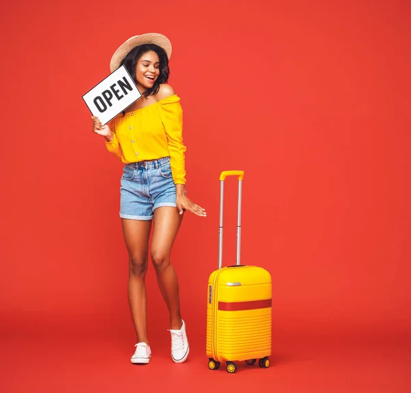 旅行的概念 快乐而兴奋的年轻黑人乐观的女性游客 提着手提箱和开着的标志 微笑着 望着红色的背风 — 图库照片