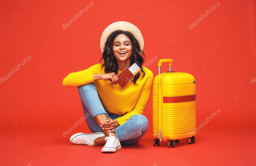 Full body positive ethnic woman with passport and tickets smiling and looking at camera while sitting near luggage during summer vacation against yellow backgroun